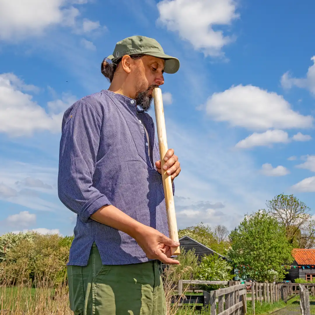 A man in profile playing a flute made from a branch with blue sky and fen landscape in background