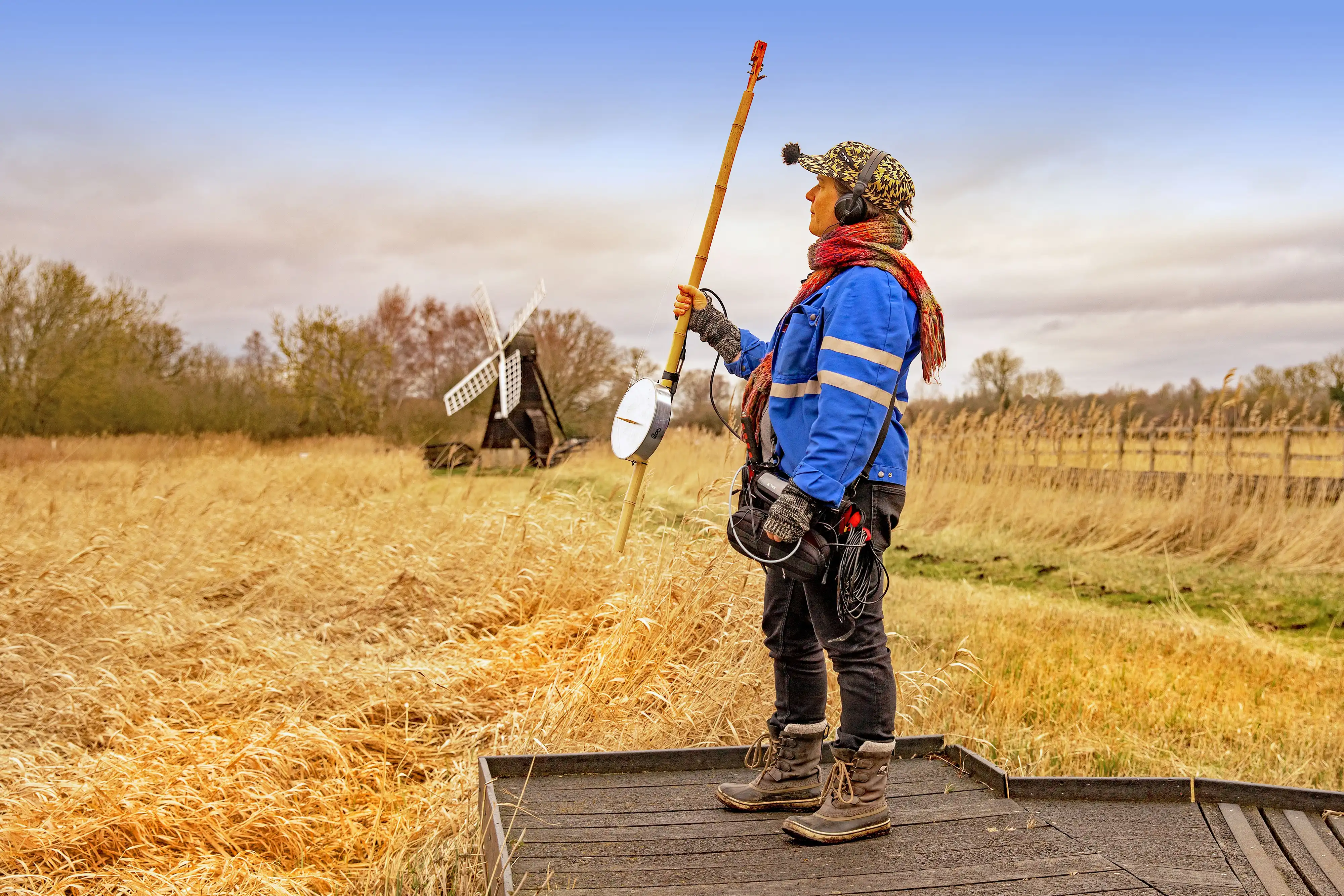 person standing holding an aeolian harp up into the oncoming wind next to a reed bed with wind pump in background