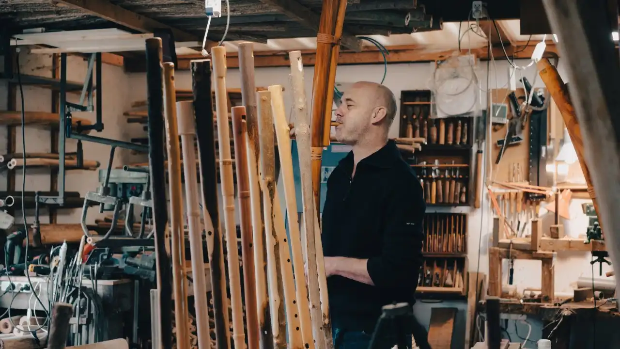 A white man wearing a black shirt playing a tall fujara flute that is clearly made from a branch, in a workshop that is full of flutes made from branches
